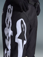 Alpinestars 2026 Racer Air Riway Pants, pantalones de motocross negros con un llamativo logotipo blanco de Alpinestars en el muslo, primer plano que muestra el tejido técnico y la correa ajustable de la cintura, usados con guantes negros para conducción off-road