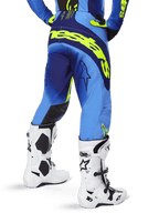 Pantalones Alpinestars Techstar Melt 2025, pantalones de motocross, azul y amarillo flúor, vista trasera que muestra un gran logotipo amarillo en el asiento y los muslos, usados con botas de motocross blancas y jersey azul a juego, ajuste atlético de competición para conducción off-road