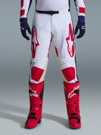 Alpinestars 2026 Racer Riway Pants, pantalones de motocross, blanco, azul y rojo, usados con botas rojas y guantes azul marino, con grandes logotipos rojos en los muslos y paneles rojos en las rodillas, diseño deportivo precurvado con hebilla de cintura ajustable