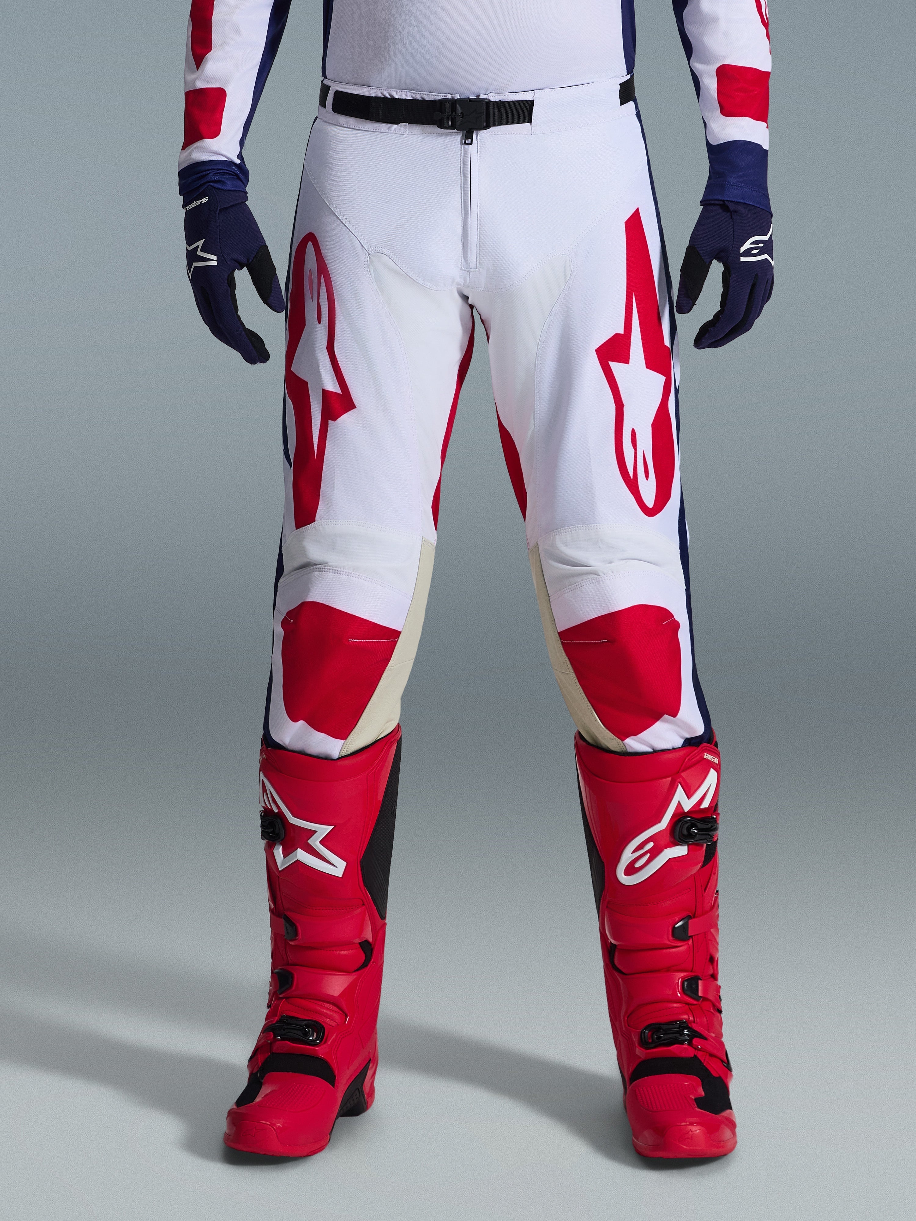 Alpinestars 2026 Racer Riway Pants, pantalones de motocross, blanco, azul y rojo, usados con botas rojas y guantes azul marino, con grandes logotipos rojos en los muslos y paneles rojos en las rodillas, diseño deportivo precurvado con hebilla de cintura ajustable
