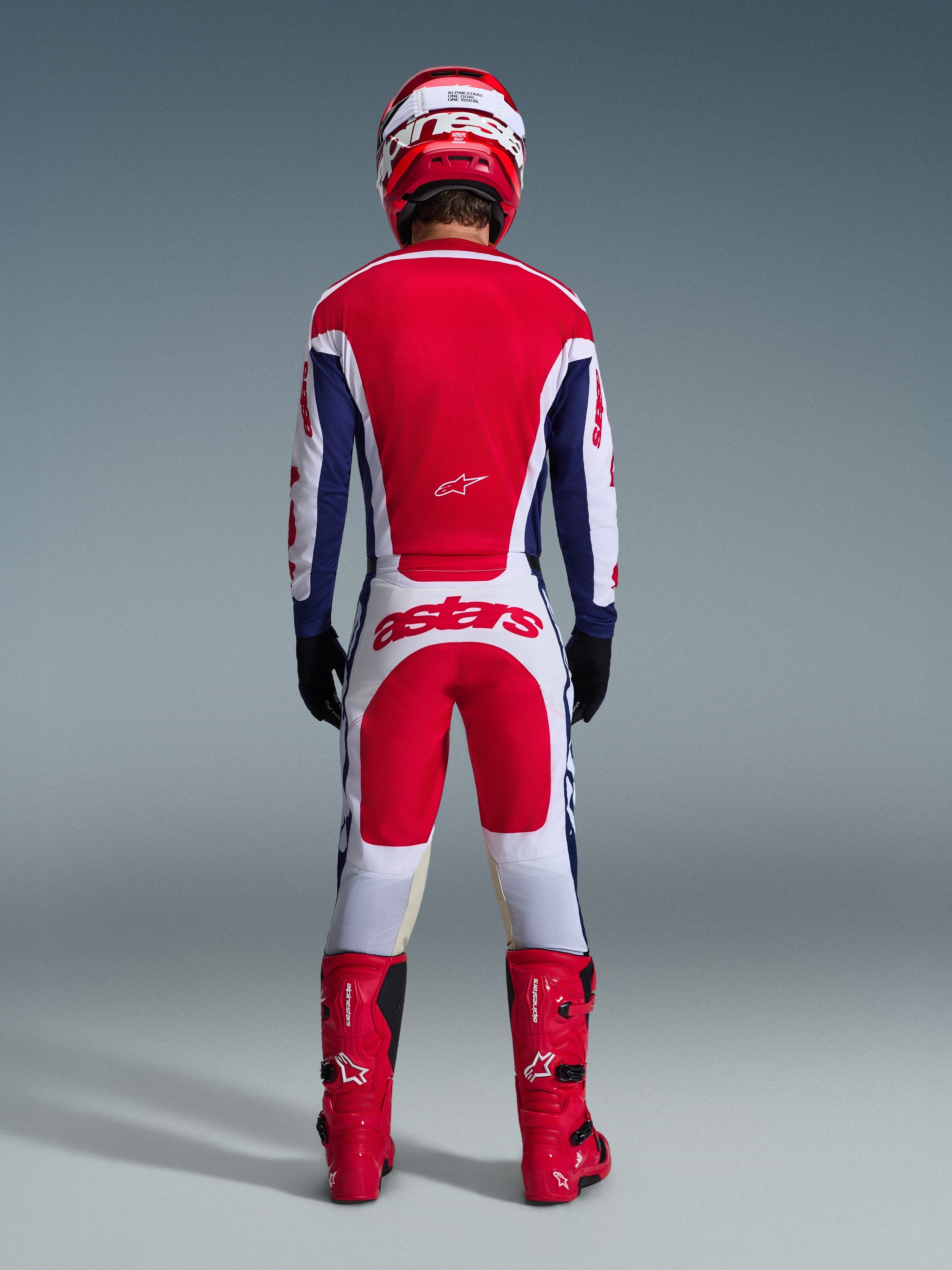 Piloto de motocross visto desde atrás con equipamiento completo de carreras, pantalones Alpinestars Racer Riway 2026 en blanco, azul y rojo con jersey a juego, botas y casco rojos, con un gran logotipo 'astars' en la parte trasera, conjunto de motocross coordinado