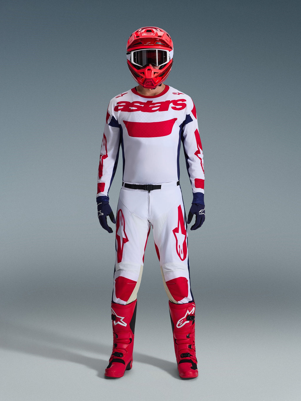 Piloto de motocross con equipamiento completo de competición, jersey blanco con marca Alpinestars en rojo y detalles azules, pantalones Alpinestars 2026 Racer Riway en blanco, azul y rojo, casco rojo con gafas a juego, botas rojas y negras, guantes azul marino, conjunto coordinado para carreras off-road