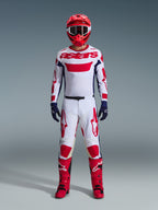 Piloto de motocross con el conjunto Alpinestars 2026 Racer Riway, jersey blanco con marca "astars" roja y paneles laterales azules, pantalones blancos con gráficos rojos, casco y botas de carreras rojas, guantes azul marino, equipamiento completo y coordinado para carreras de motocross