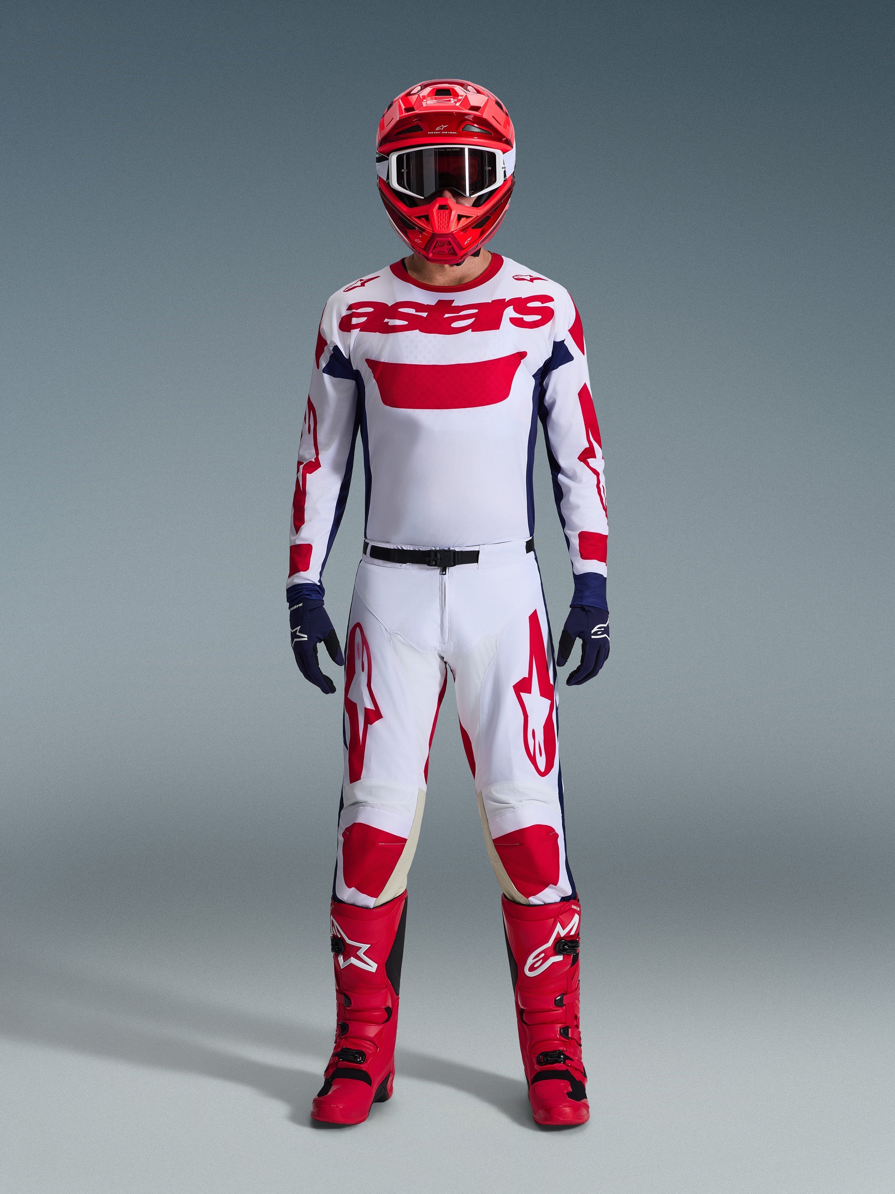 Motocross rider wearing the Alpinestars 2026 Racer Riway Gear Set, White jersey with red "astars" branding and blue side panels, white pants with red graphics, red racing helmet and boots, navy blue gloves, complete coordinated motocross racing outfit