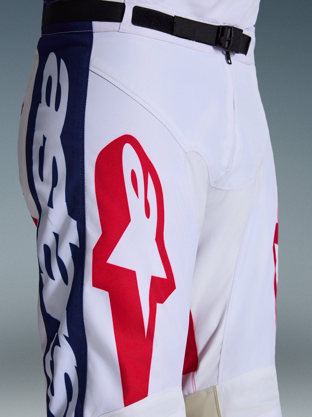 Alpinestars 2026 Racer Riway Pants, pantalones de motocross, blanco, azul y rojo, primer plano de la parte superior de la pierna y la cintura que muestra un gran logo de estrella roja sobre tejido técnico blanco, paneles laterales azules con marca y cinturón ajustable negro con hebilla magnética SX2, diseño deportivo de carreras off-road