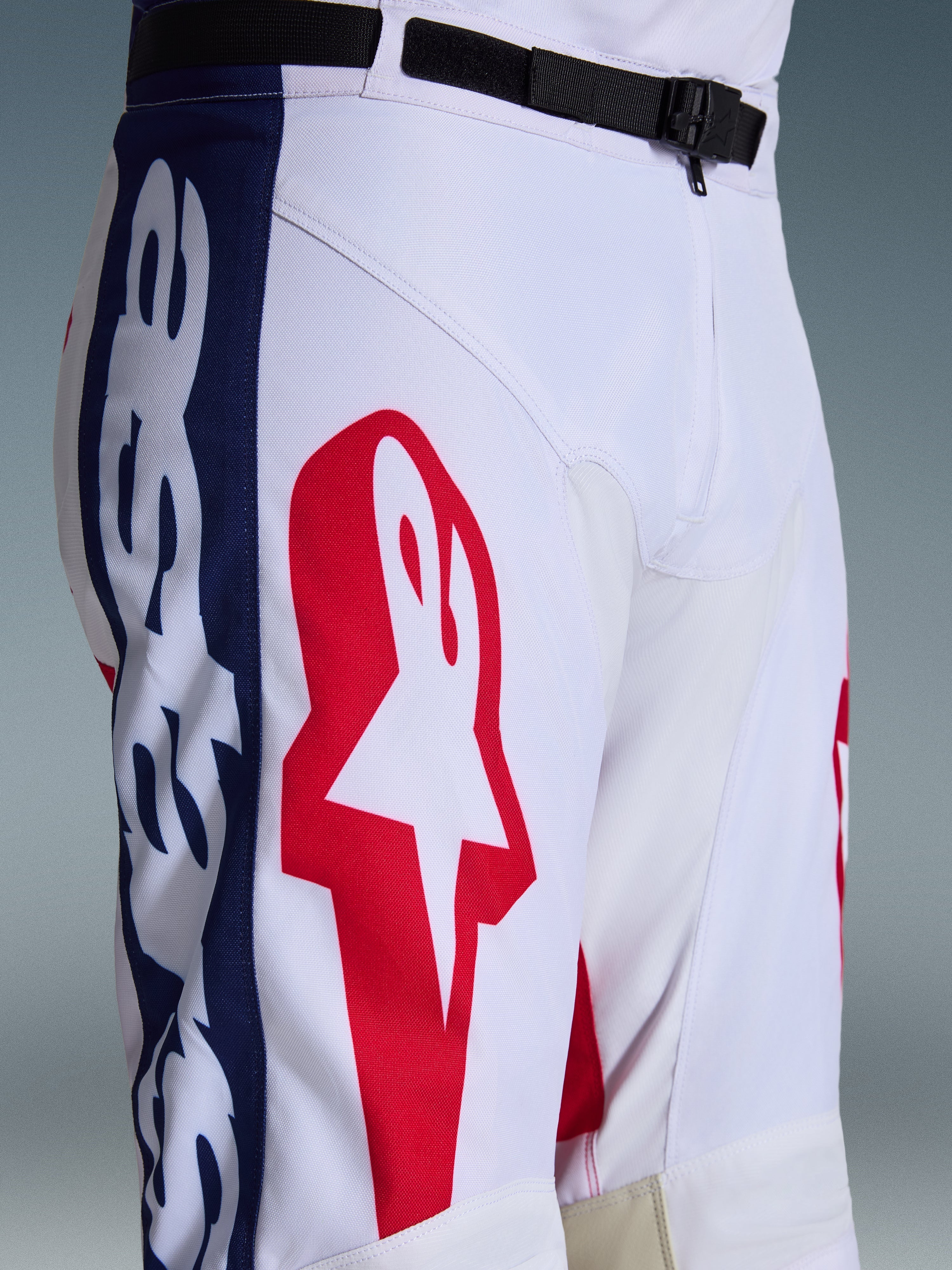 Alpinestars 2026 Racer Riway Pants, pantalones de motocross, blanco, azul y rojo, primer plano de la parte superior de la pierna y la cintura que muestra un gran logo de estrella roja sobre tejido técnico blanco, paneles laterales azules con marca y cinturón ajustable negro con hebilla magnética SX2, diseño deportivo de carreras off-road