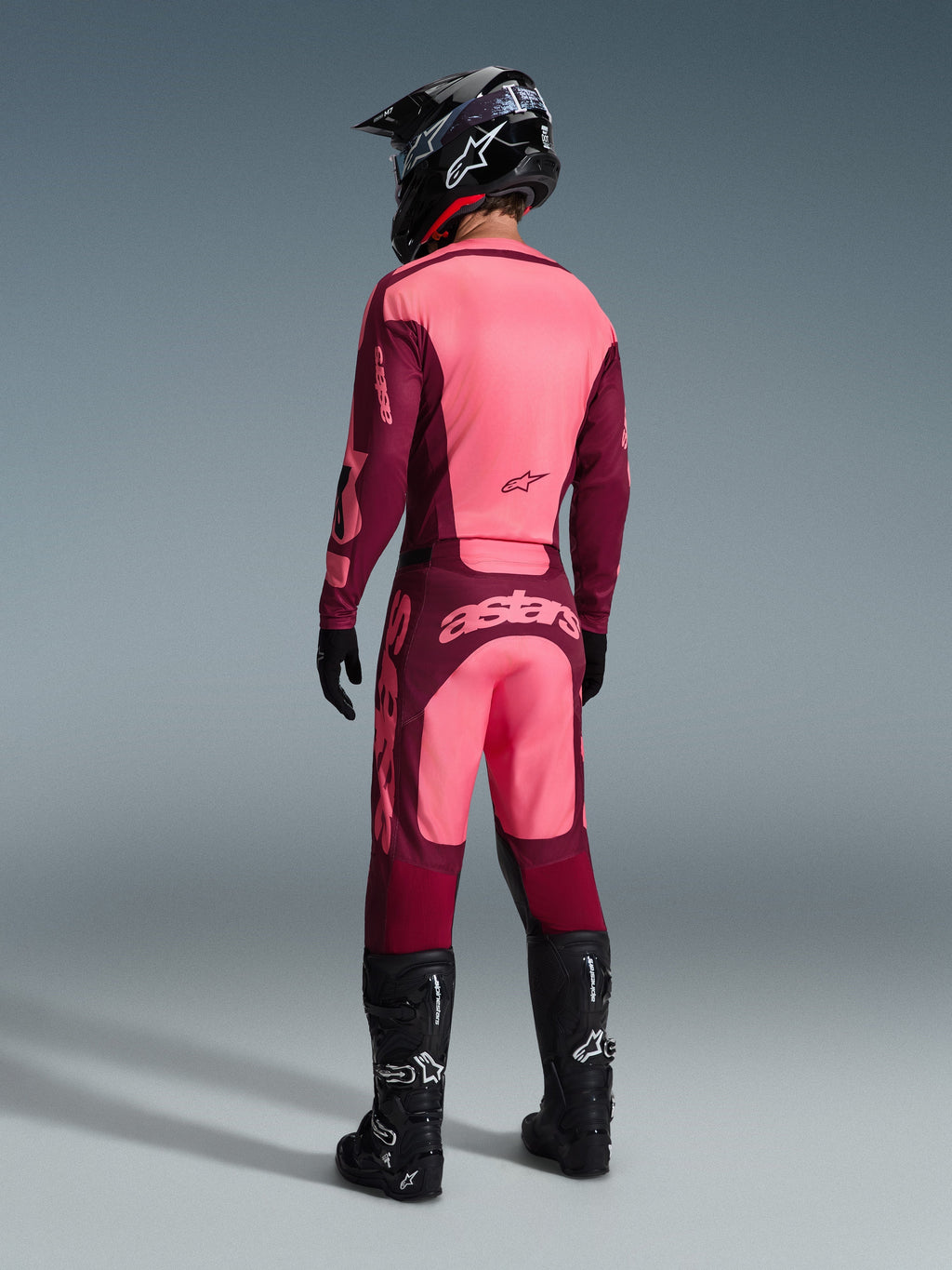 Alpinestars 2026 Racer Riway Jersey and matching pants, Motocross gear in Dark Red Coral, rear view of rider wearing black helmet and black boots, featuring vibrant coral and maroon panels with bold Astars branding on the back