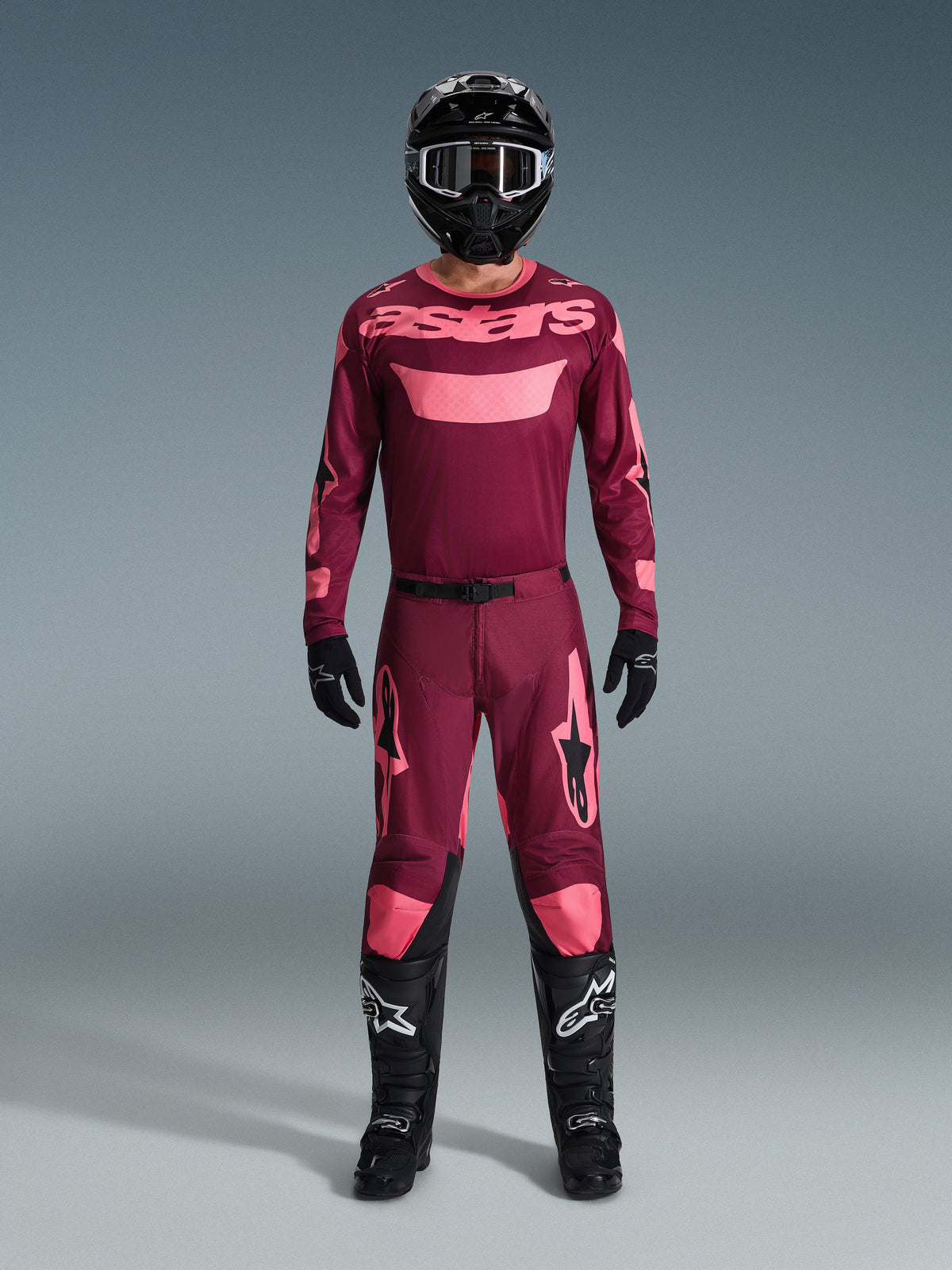 Piloto de motocross luciendo el conjunto Alpinestars 2026 Racer Riway, camiseta granate con logotipos 'astars' en rosa, pantalones granates a juego con detalles rosas en las rodillas y las piernas, casco negro, guantes negros y botas negras, equipamiento de alto rendimiento completo para carreras off-road