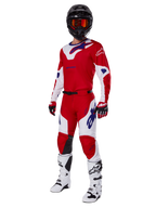 Alpinestars 2025 Racer Veil Pants, pantalones de motocross, rojo brillante y blanco, usados como parte de un conjunto completo de equipamiento de competición que incluye una camiseta roja y blanca a juego, casco negro con gafas naranjas, botas blancas y guantes rojos, con logotipos de Alpinestars en un llamativo color azul.