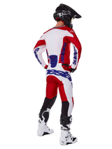 Alpinestars 2025 Racer Veil Pants, pantalones de motocross, rojo brillante y blanco con detalles azules, vistos desde atrás en un piloto con equipamiento completo, con logotipo distintivo de Alpinestars, paneles transpirables en la parte inferior de las piernas, y botas blancas y casco negro coordinados.
