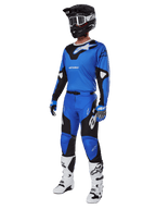 Piloto de motocross con equipamiento completo de competición, que incluye los pantalones Alpinestars 2025 Racer Veil en azul y negro, jersey azul a juego con detalles blancos, casco negro con gafas blancas, botas blancas y negras, y guantes azules, conjunto coordinado para carreras off-road
