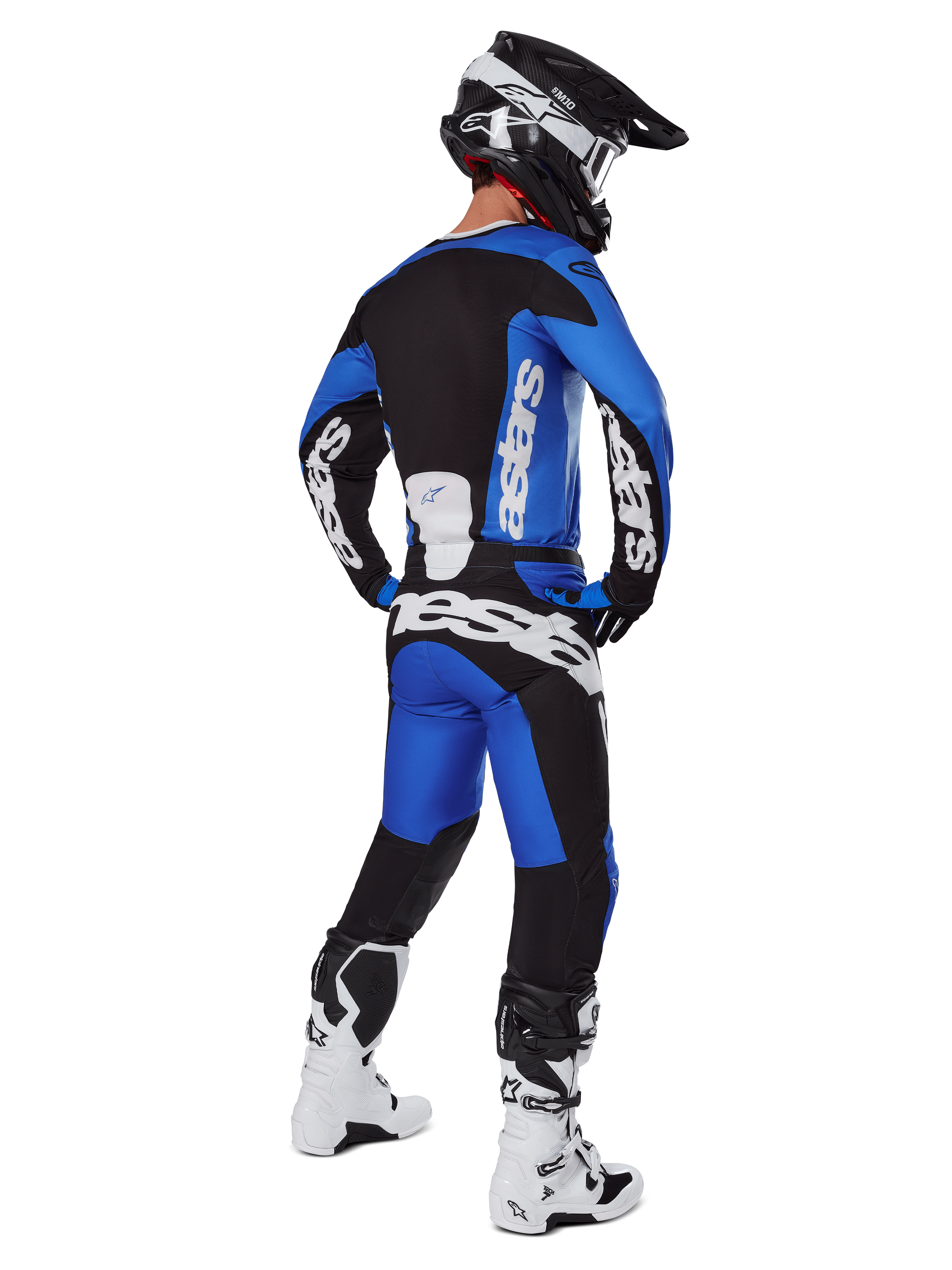 Piloto de motocross visto desde atrás con pantalones Alpinestars 2025 Racer Veil en azul y negro, jersey de manga larga a juego con logotipos blancos, botas Tech 7 en blanco y negro y casco SM10 negro, conjunto de equipamiento completo para carreras off-road