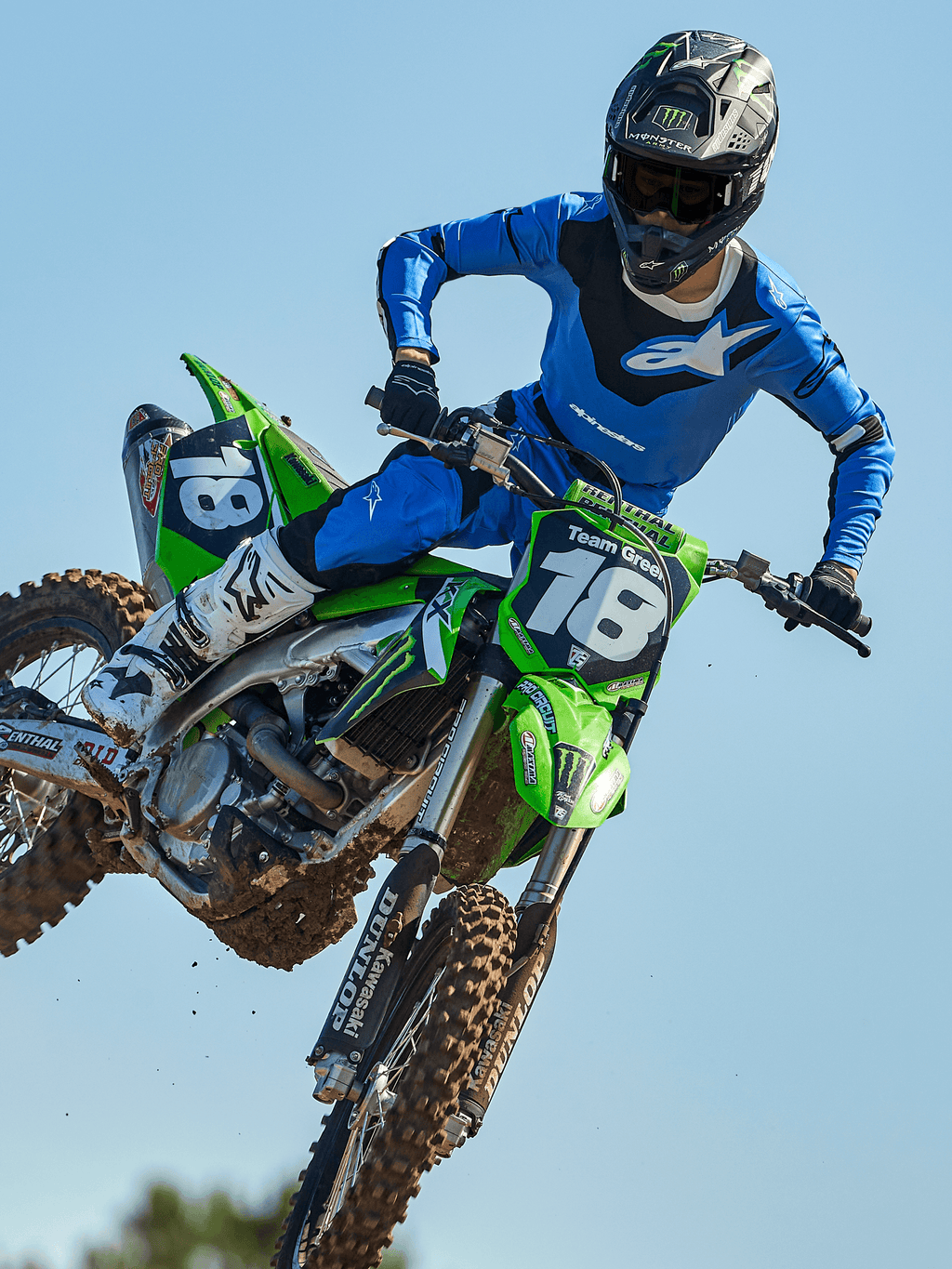 Piloto de motocross en pleno salto sobre una moto verde, con pantalones Alpinestars 2025 Racer Veil en azul y negro, jersey azul a juego con logotipo blanco en el pecho, botas blancas y negras y casco negro, equipamiento off-road de alto rendimiento para competición