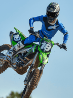 Piloto de motocross en pleno salto sobre una moto verde, con pantalones Alpinestars 2025 Racer Veil en azul y negro, jersey azul a juego con logotipo blanco en el pecho, botas blancas y negras y casco negro, equipamiento off-road de alto rendimiento para competición
