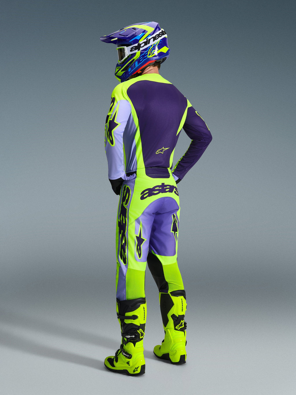 Piloto de motocross visto desde atrás con los pantalones Alpinestars 2026 Racer Portl en Purple Yellow Fluo, combinados con jersey y casco a juego en púrpura y amarillo, y botas de carreras en amarillo brillante, con el logotipo 'astars' en la parte trasera y estrellas en las piernas.