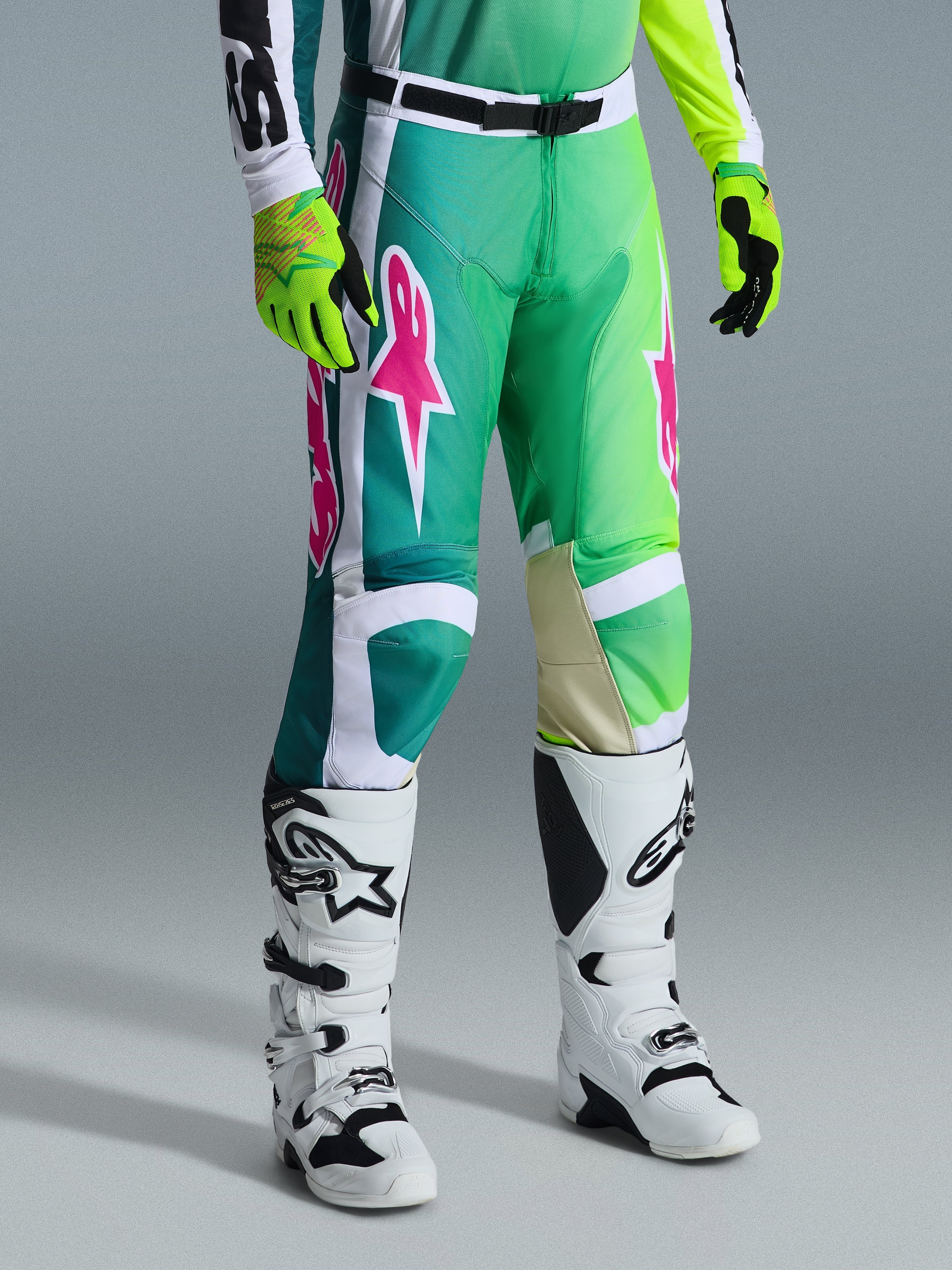 Alpinestars 2026 Racer Portl Pants, pantalones de motocross, verdes, blancos y negros con logotipos rosas, combinados con botas blancas y guantes amarillo neón, con llamativos patrones gráficos y hebilla de cintura ajustable negra, equipamiento de alto rendimiento para conducción todoterreno