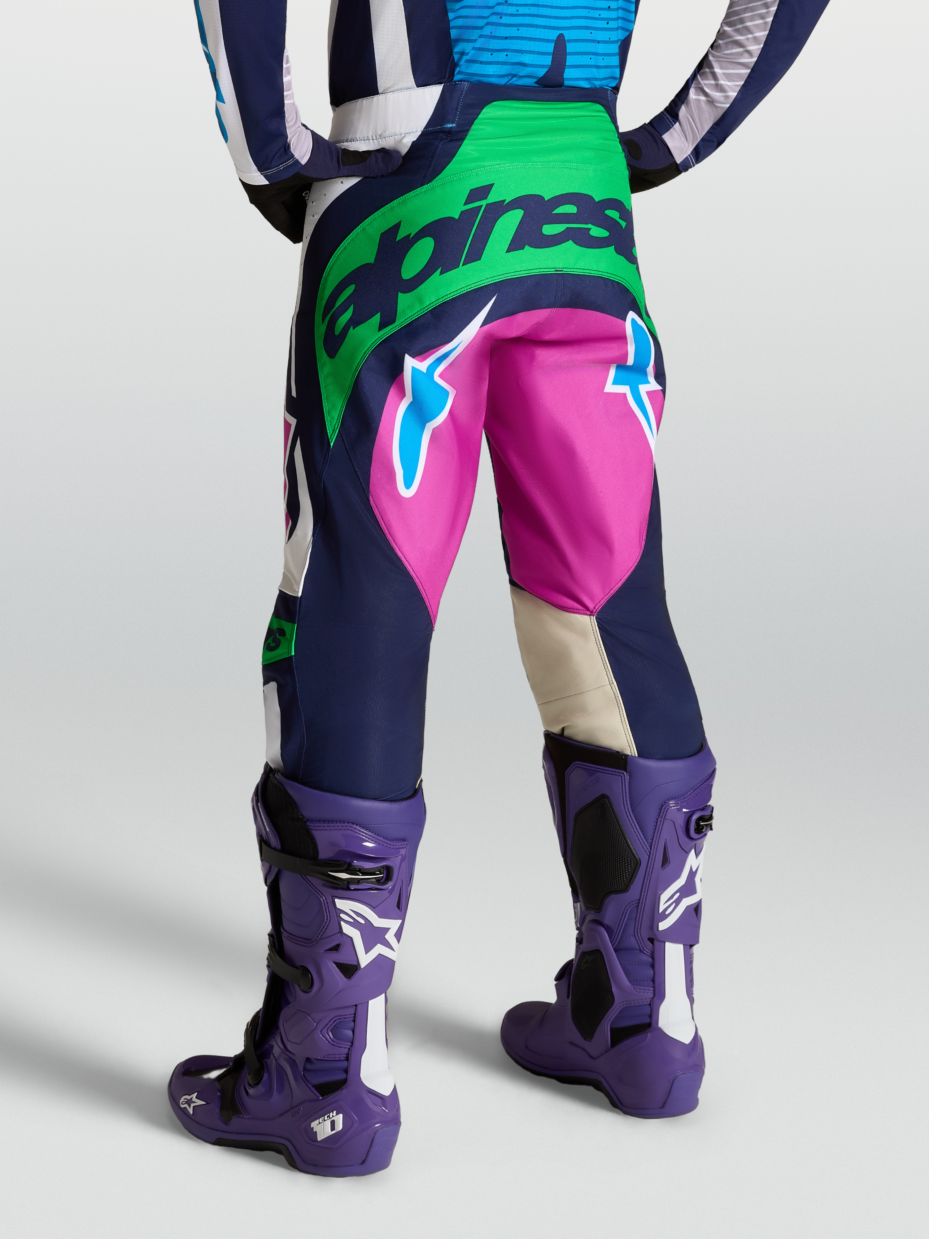Alpinestars Techstar Maven, pantalones de motocross para hombre, azul y verde con detalles en rosa, vista trasera con botas Tech 10 moradas