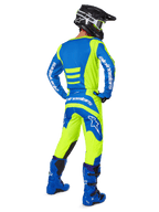 Piloto de motocross visto desde atrás vistiendo los pantalones Alpinestars 2025 Fluid Haul y el jersey a juego en amarillo flúor y azul, coordinado con botas azules y un casco Alpinestars negro, equipamiento profesional de carreras todoterreno
