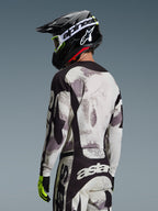 Piloto de motocross desde atrás con el Jersey Racer Lahnd 2026 en Sand Camo, jersey con patrón abstracto gris y blanco con detalles negros, combinado con casco Alpinestars negro, gafas blancas y guantes amarillo neón, mostrando el perfil trasero y el ajuste técnico para competición off-road