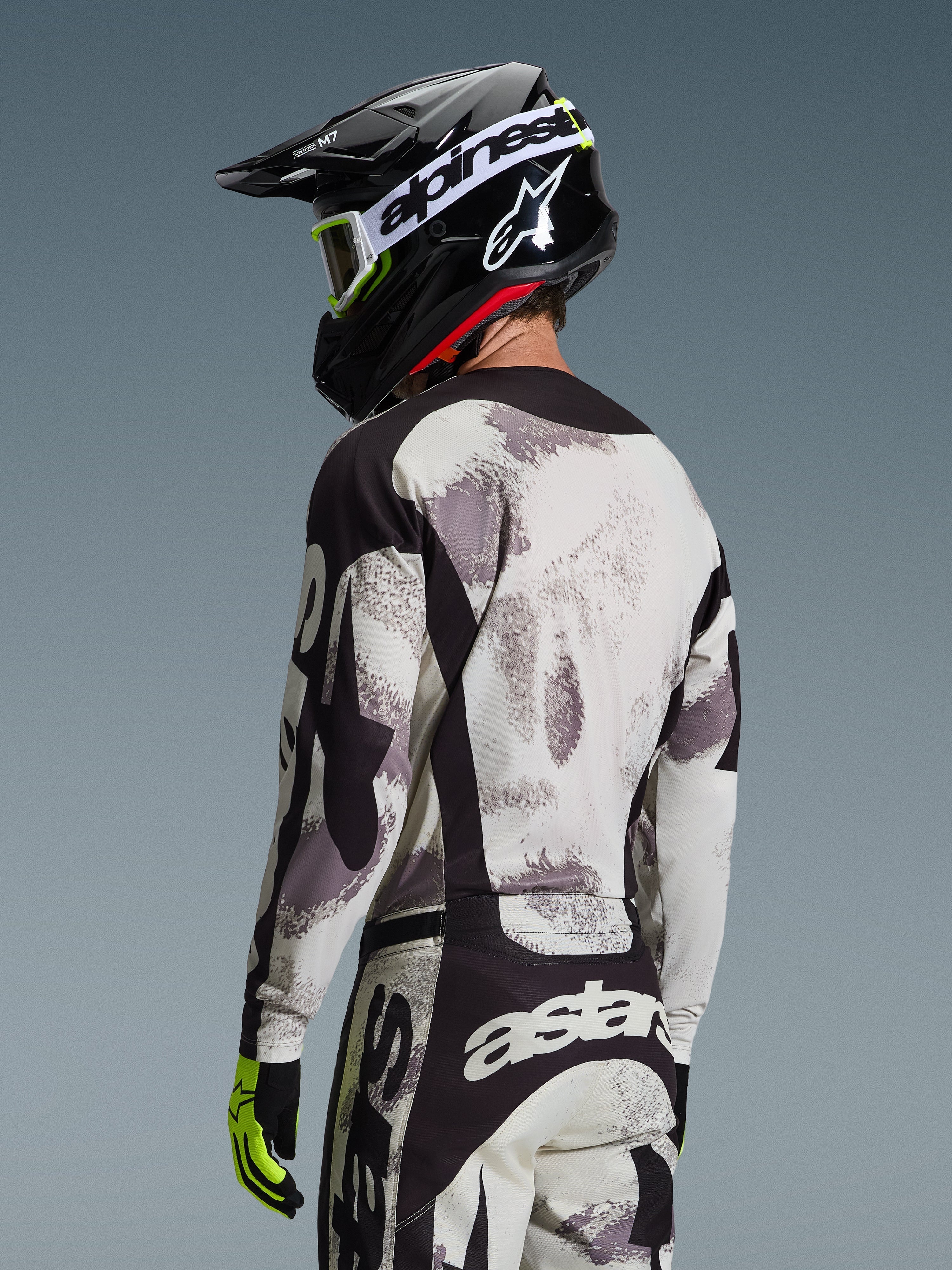 Piloto de motocross desde atrás con el Jersey Racer Lahnd 2026 en Sand Camo, jersey con patrón abstracto gris y blanco con detalles negros, combinado con casco Alpinestars negro, gafas blancas y guantes amarillo neón, mostrando el perfil trasero y el ajuste técnico para competición off-road