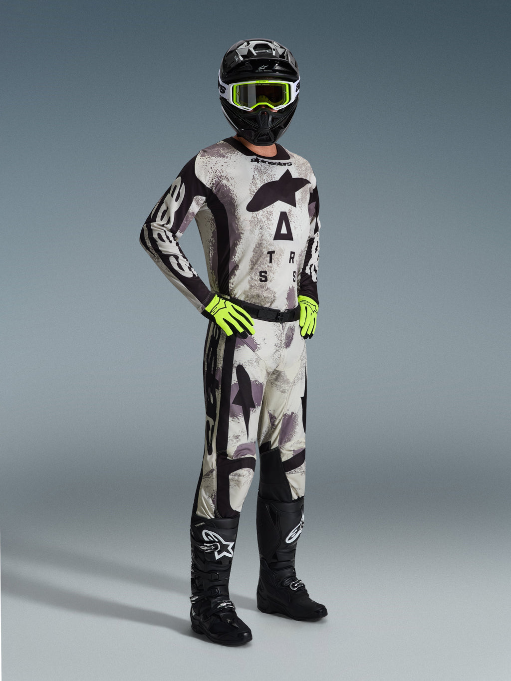 Piloto de motocross con el Jersey Racer Lahnd 2026 en Sand Camo, patrón de camuflaje gris y negro con marca Alpinestars, pantalones de motocross a juego, guantes amarillo neón, casco negro con gafas amarillas y botas negras, equipamiento profesional completo para carreras off-road.