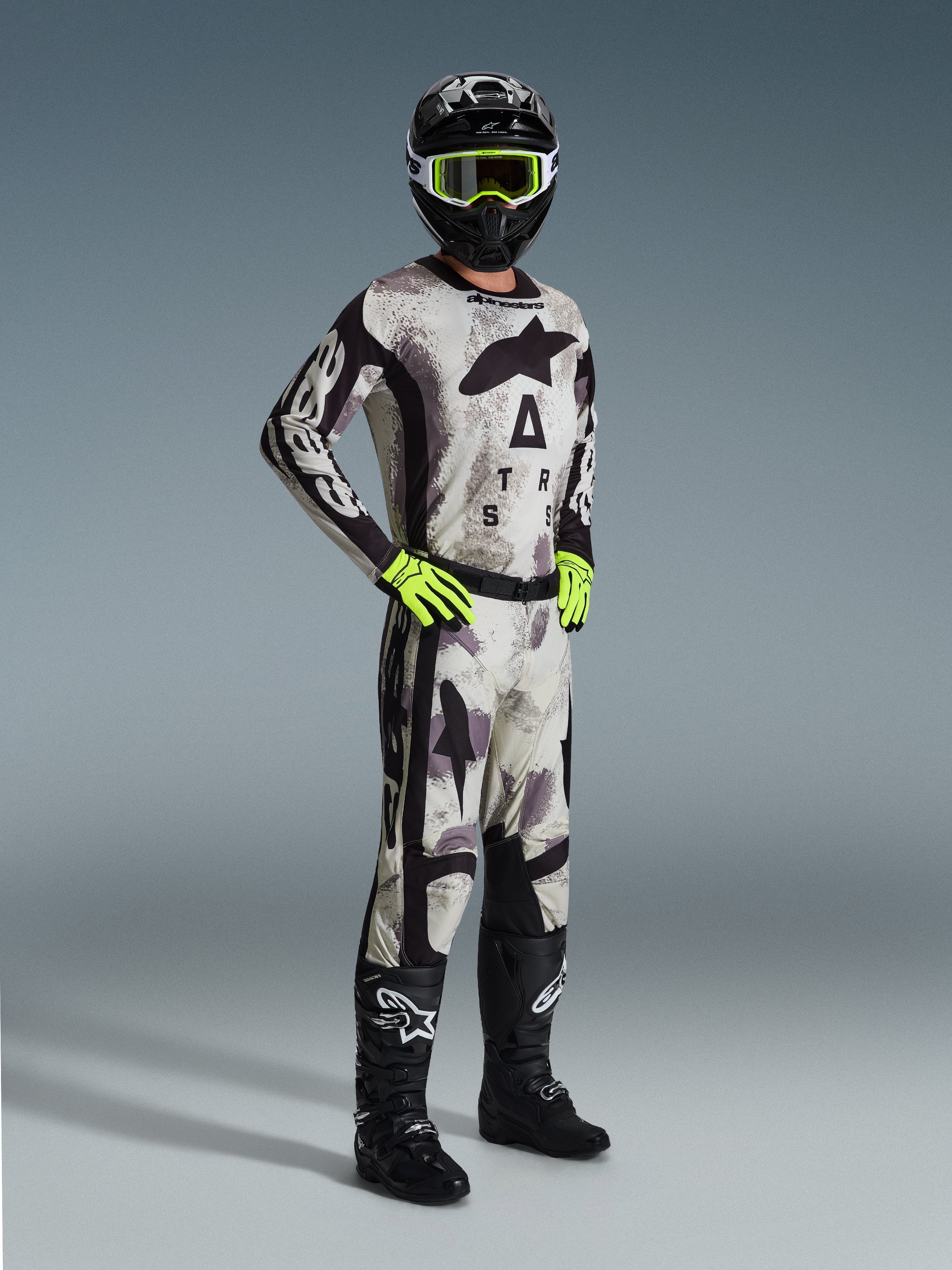 Piloto de motocross con el Jersey Racer Lahnd 2026 en Sand Camo, patrón de camuflaje gris y negro con marca Alpinestars, pantalones de motocross a juego, guantes amarillo neón, casco negro con gafas amarillas y botas negras, equipamiento profesional completo para carreras off-road.