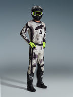 Piloto de motocross con equipamiento completo de Alpinestars, pantalones 2026 Racer Lahnd en Sand Camo, jersey de camuflaje a juego con logotipos llamativos, casco negro con gafas amarillo neón, guantes verde neón y botas negras, conjunto profesional para rendimiento off-road.
