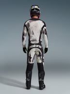 Motocross rider wearing complete Alpinestars 2026 Racer Lahnd Gear Set, viewed from behind, Sand Camo jersey and pants with light gray and black abstract pattern, black helmet, black and neon yellow gloves, black boots, featuring 'astars' branding on the rear of the pants