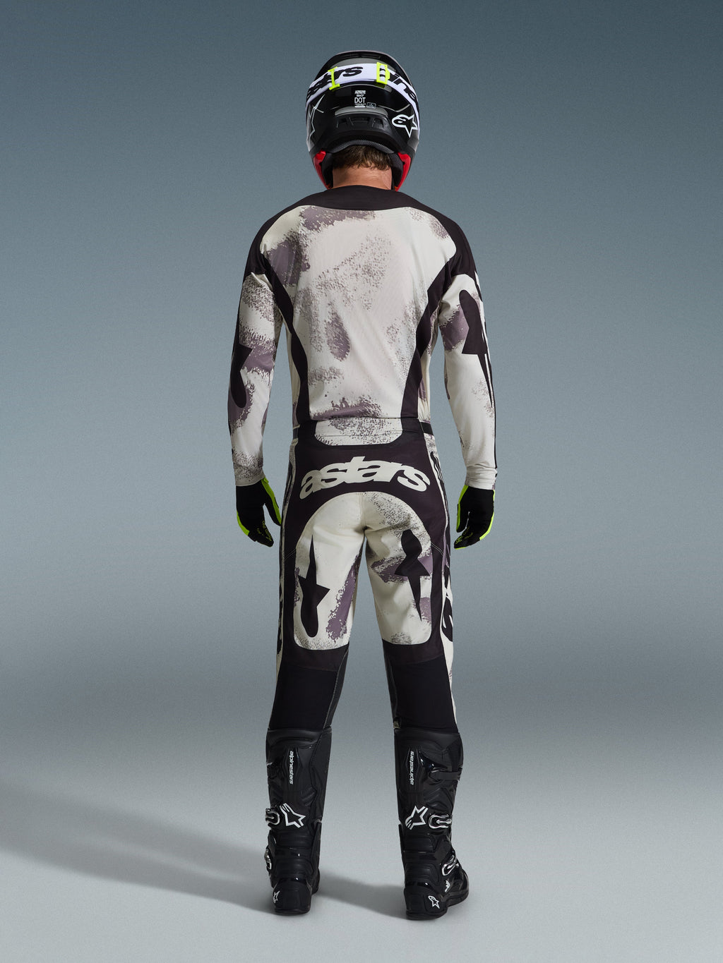 Piloto de motocross visto desde atrás con el jersey Racer Lahnd 2026 y pantalones a juego en color Sand Camo, con un patrón abstracto crema y gris y paneles negros, marca 'astars' en los pantalones, botas Alpinestars negras y casco negro para un look completo de carreras off-road.