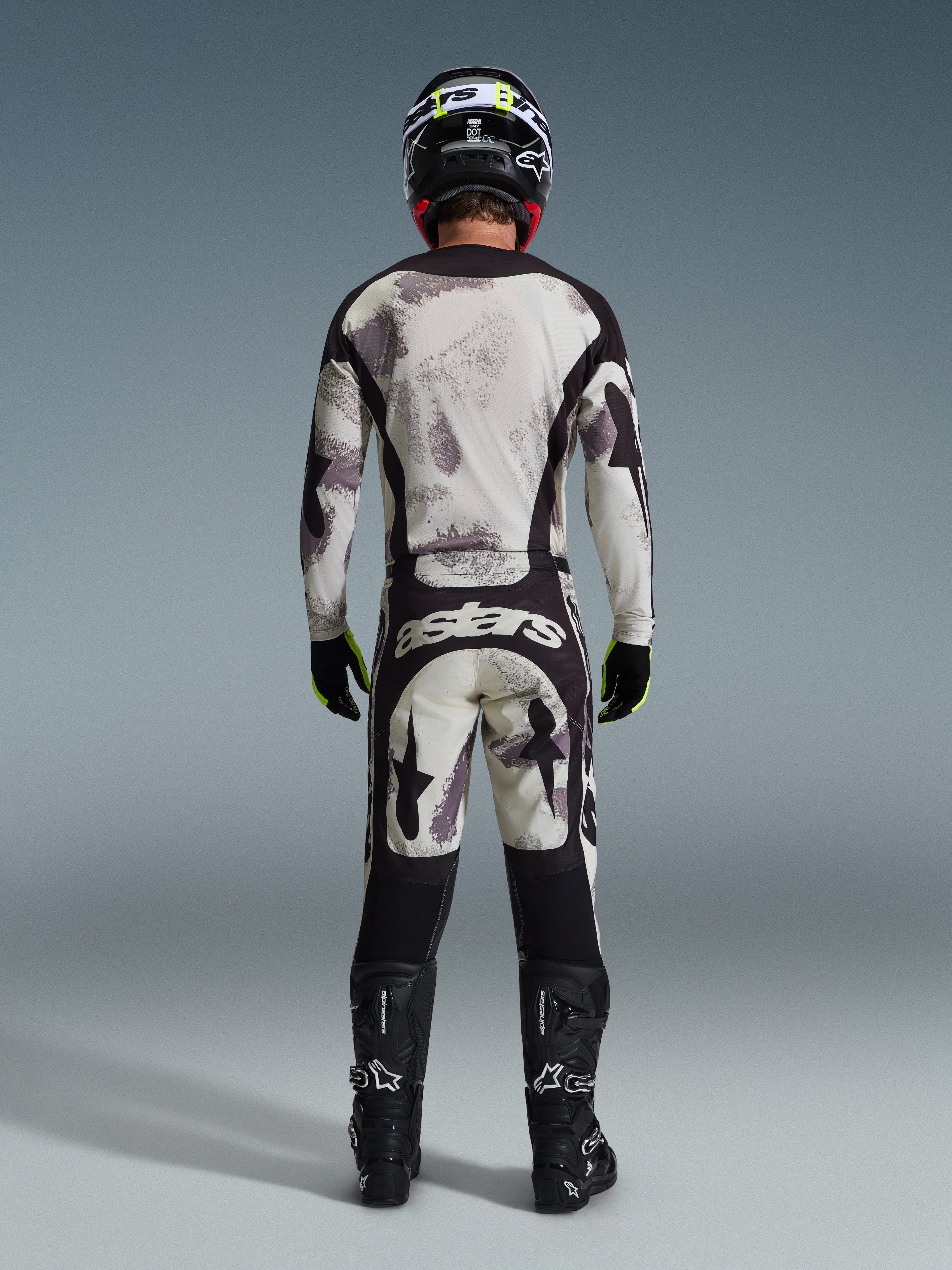Piloto de motocross visto desde atrás con el jersey Racer Lahnd 2026 y pantalones a juego en color Sand Camo, con un patrón abstracto crema y gris y paneles negros, marca 'astars' en los pantalones, botas Alpinestars negras y casco negro para un look completo de carreras off-road.