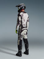 Piloto de motocross con equipamiento completo, Jersey Racer Lahnd 2026 y pantalones a juego en Sand Camo de Alpinestars, patrón de camuflaje gris y negro con logotipos destacados, casco negro, botas negras y guantes amarillo neón, vista de perfil trasera