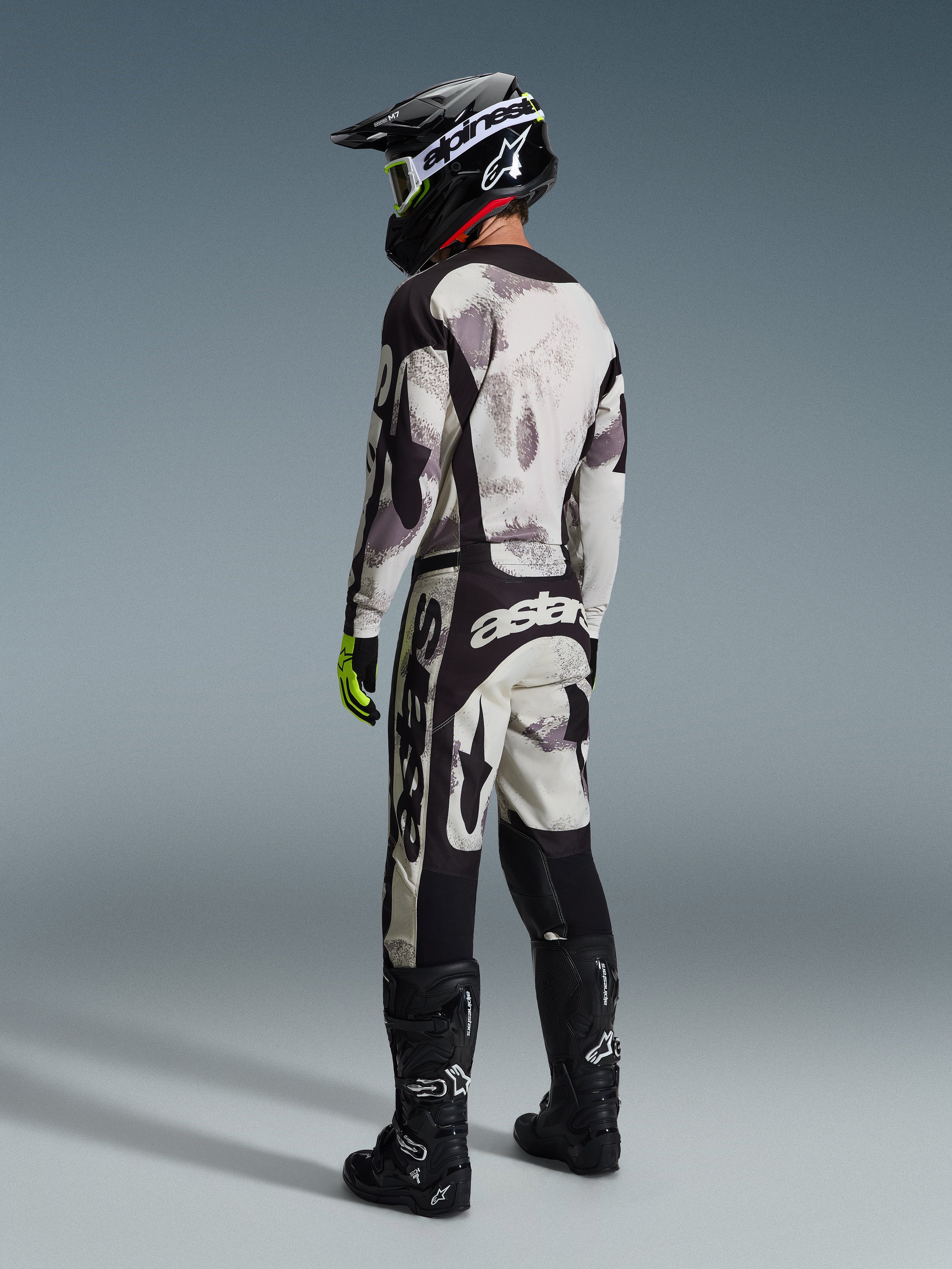 Piloto de motocross con equipamiento completo, Jersey Racer Lahnd 2026 y pantalones a juego en Sand Camo de Alpinestars, patrón de camuflaje gris y negro con logotipos destacados, casco negro, botas negras y guantes amarillo neón, vista de perfil trasera