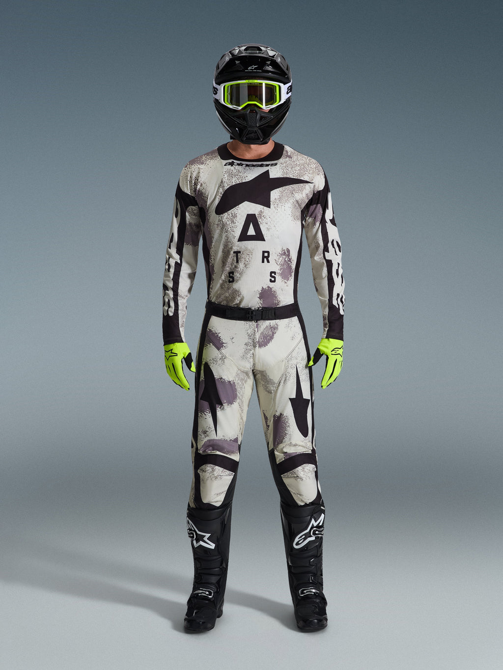 Piloto de motocross con equipamiento de competición completo, pantalones Alpinestars Racer Lahnd 2026 y jersey a juego en gris Sand Camo, casco negro con gafas amarillo neón, guantes amarillo neón brillante y botas negras, conjunto profesional coordinado para carreras off-road