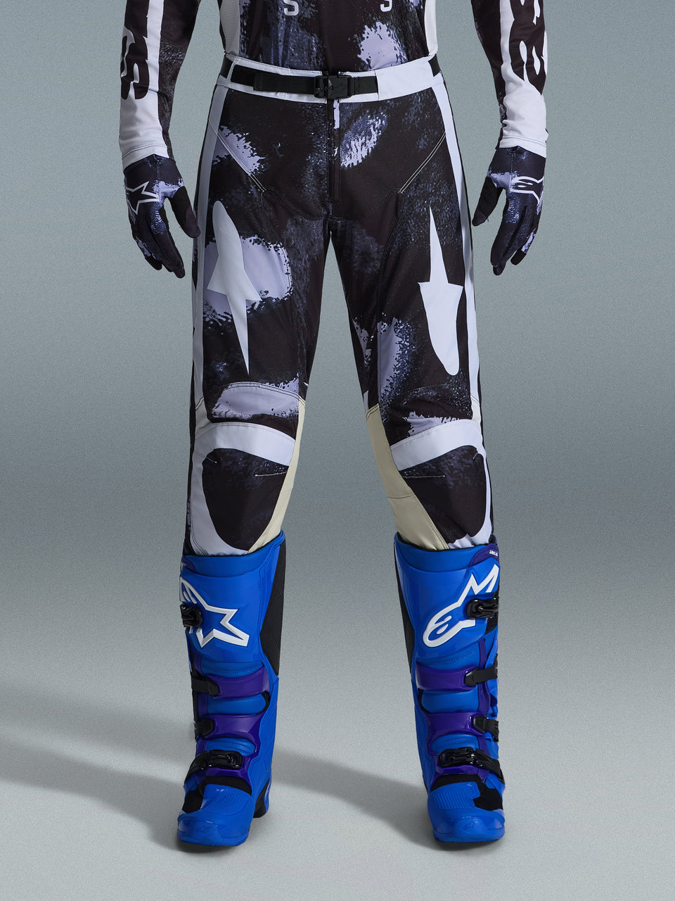 Alpinestars 2026 Racer Lahnd Pants, Motocross Pants, Iron Camo, worn with matching camouflage gloves and bright blue and purple boots, featuring stylized white star graphics on a dark camo pattern, technical magnetic buckle closure, professional off-road racing gear.