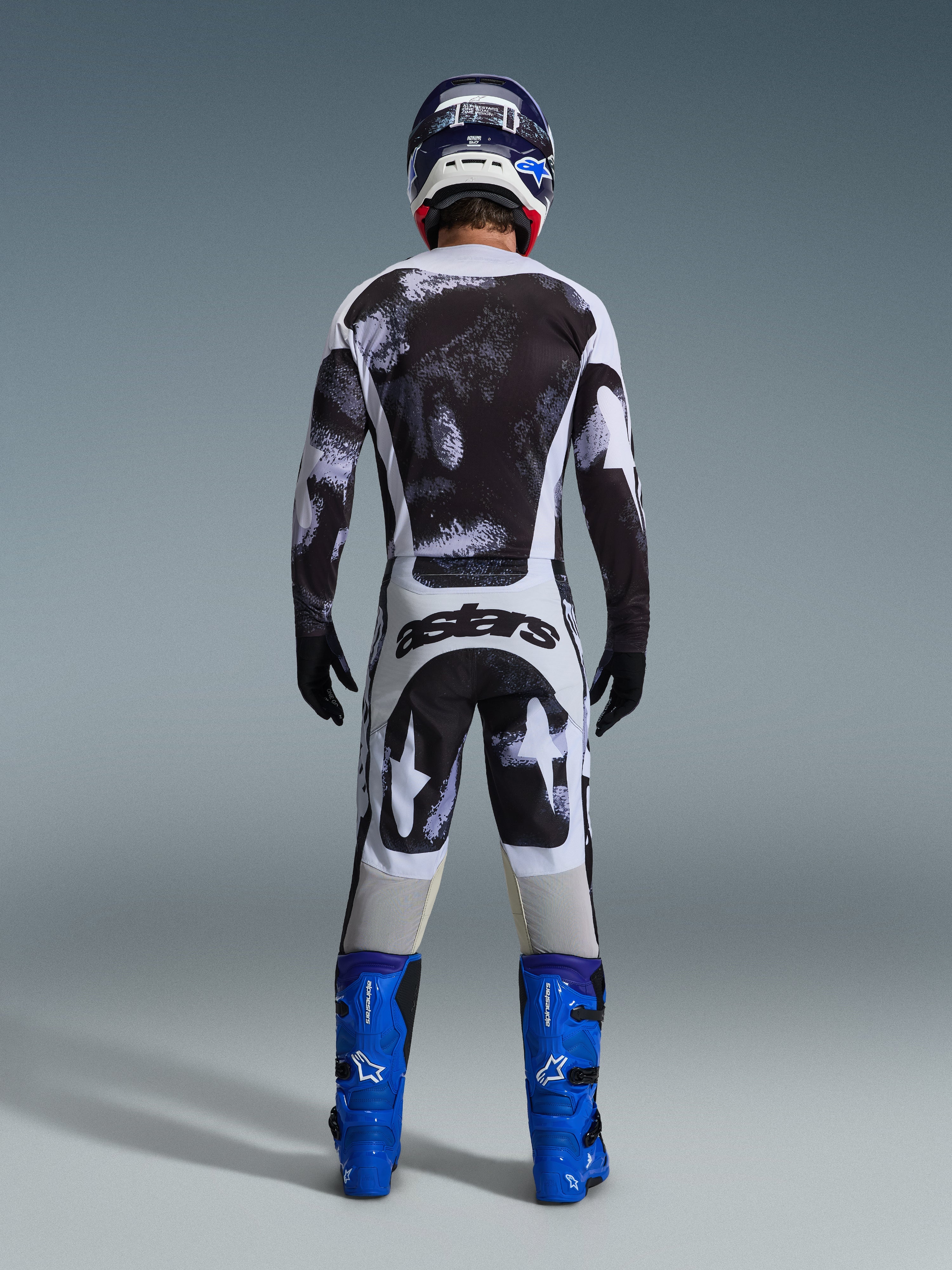 Motocross rider from behind wearing the Alpinestars 2026 Racer Lahnd Jersey and matching pants in Iron Camo, featuring a black and white splatter pattern, paired with bright blue boots and a coordinating helmet, complete gear set for off-road racing