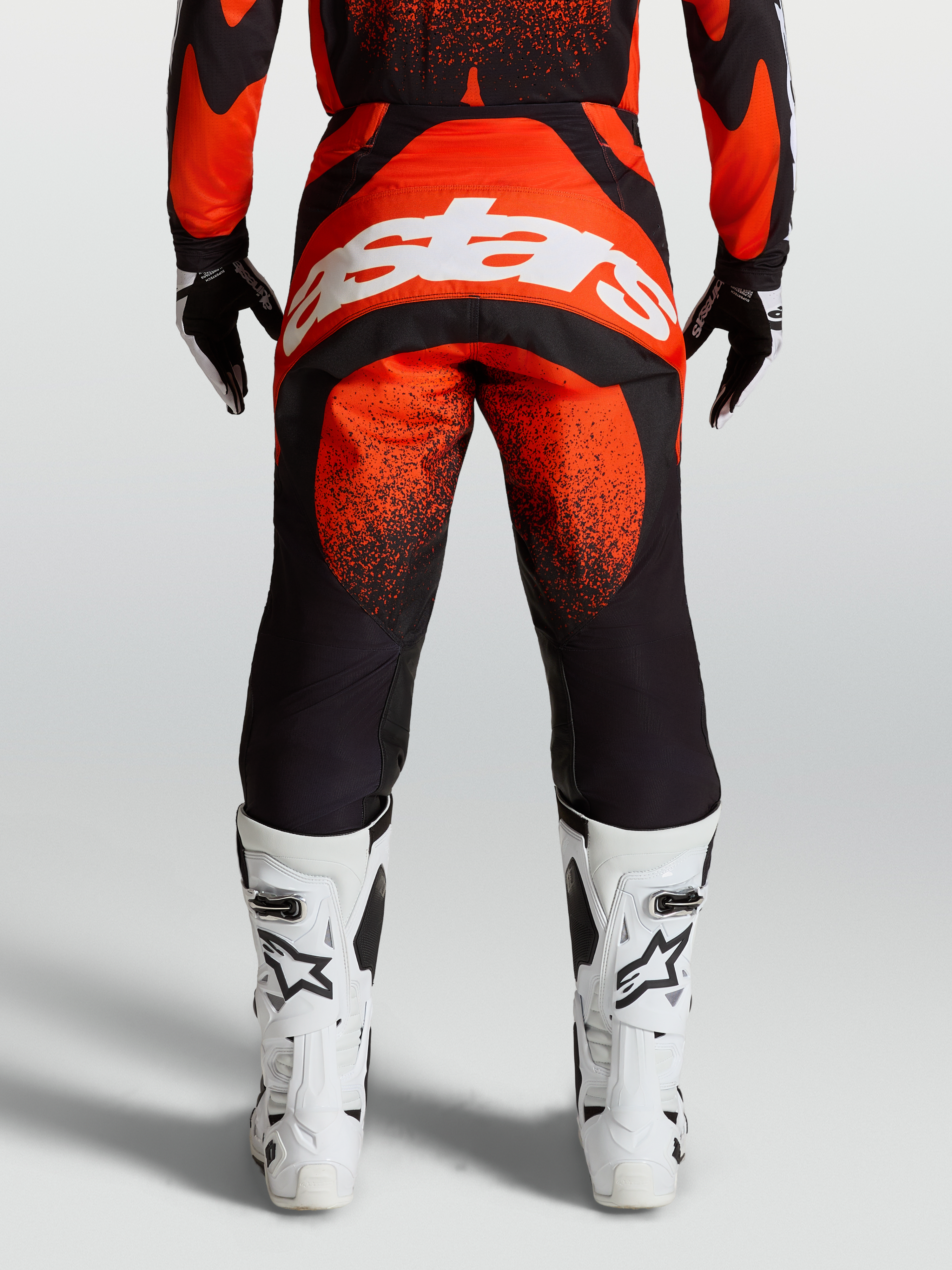 Alpinestars Techstar Swan, pantalones de motocross para hombre, rojos y negros con logo blanco en el asiento, vista trasera en modelo con botas blancas de carreras