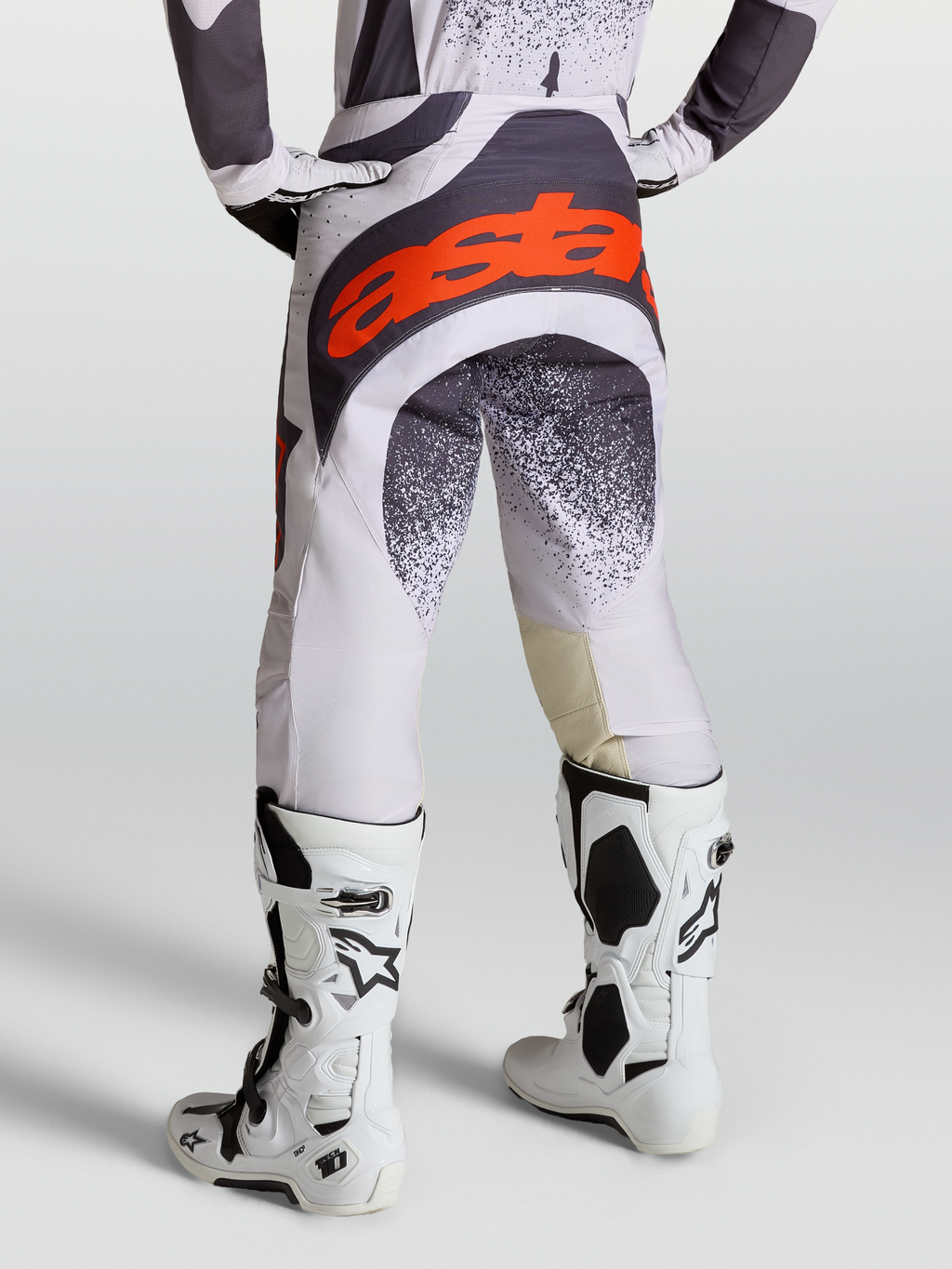 Alpinestars Pantalones Techstar Swan, pantalones de motocross para hombre, lila claro y gris oscuro con detalles en rojo flúor, usados con botas Tech 10 blancas.