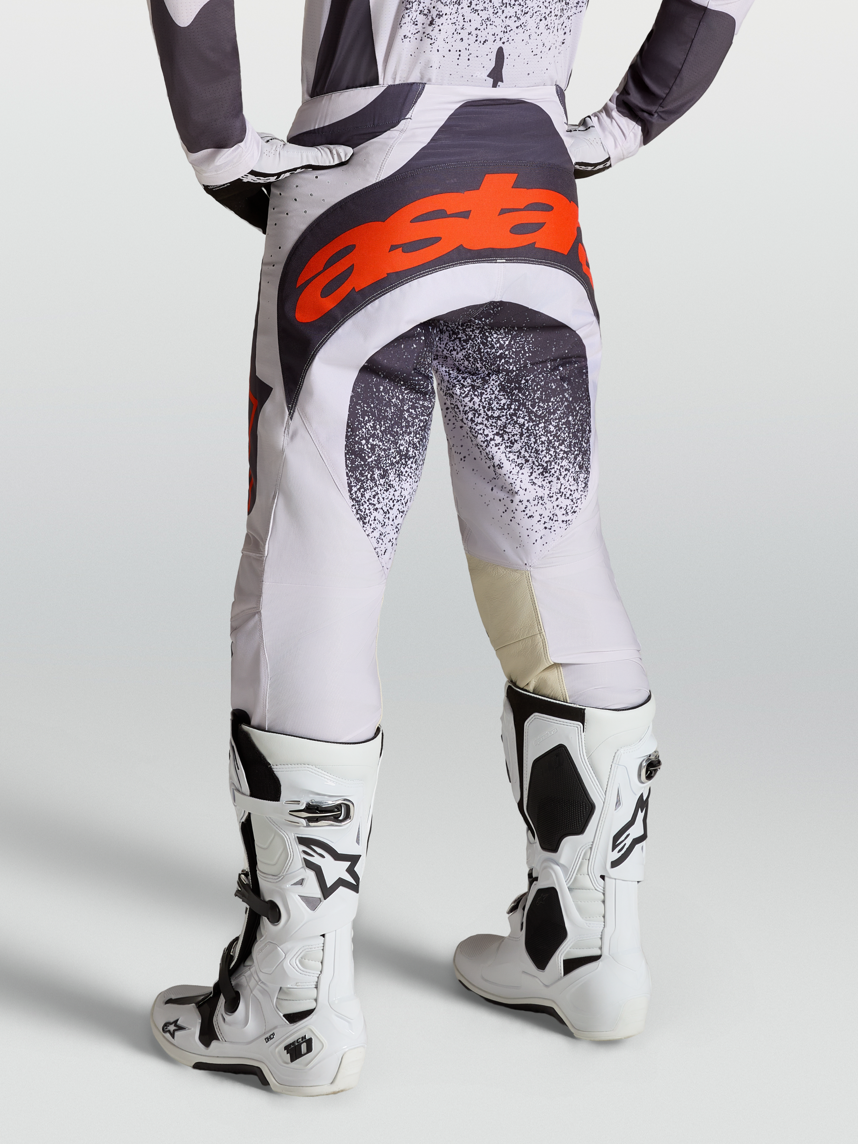Alpinestars Pantalones Techstar Swan, pantalones de motocross para hombre, lila claro y gris oscuro con detalles en rojo flúor, usados con botas Tech 10 blancas.