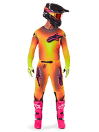 Piloto de motocross con la equipación Alpinestars 2025 Supertech Lipan, con un vibrante diseño degradado en amarillo y rosa, jersey de manga larga a juego, botas rosa brillante, guantes verde lima y casco negro con gafas naranjas.