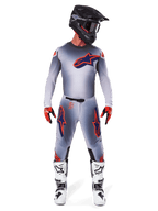 Piloto de motocross con pantalones Alpinestars Supertech Lipan 2025 y jersey a juego en Gris Naranja Flúor, con logotipos llamativos en naranja y azul marino, casco negro, guantes rojos y botas blancas y negras, kit de competición completo para rendimiento todoterreno.