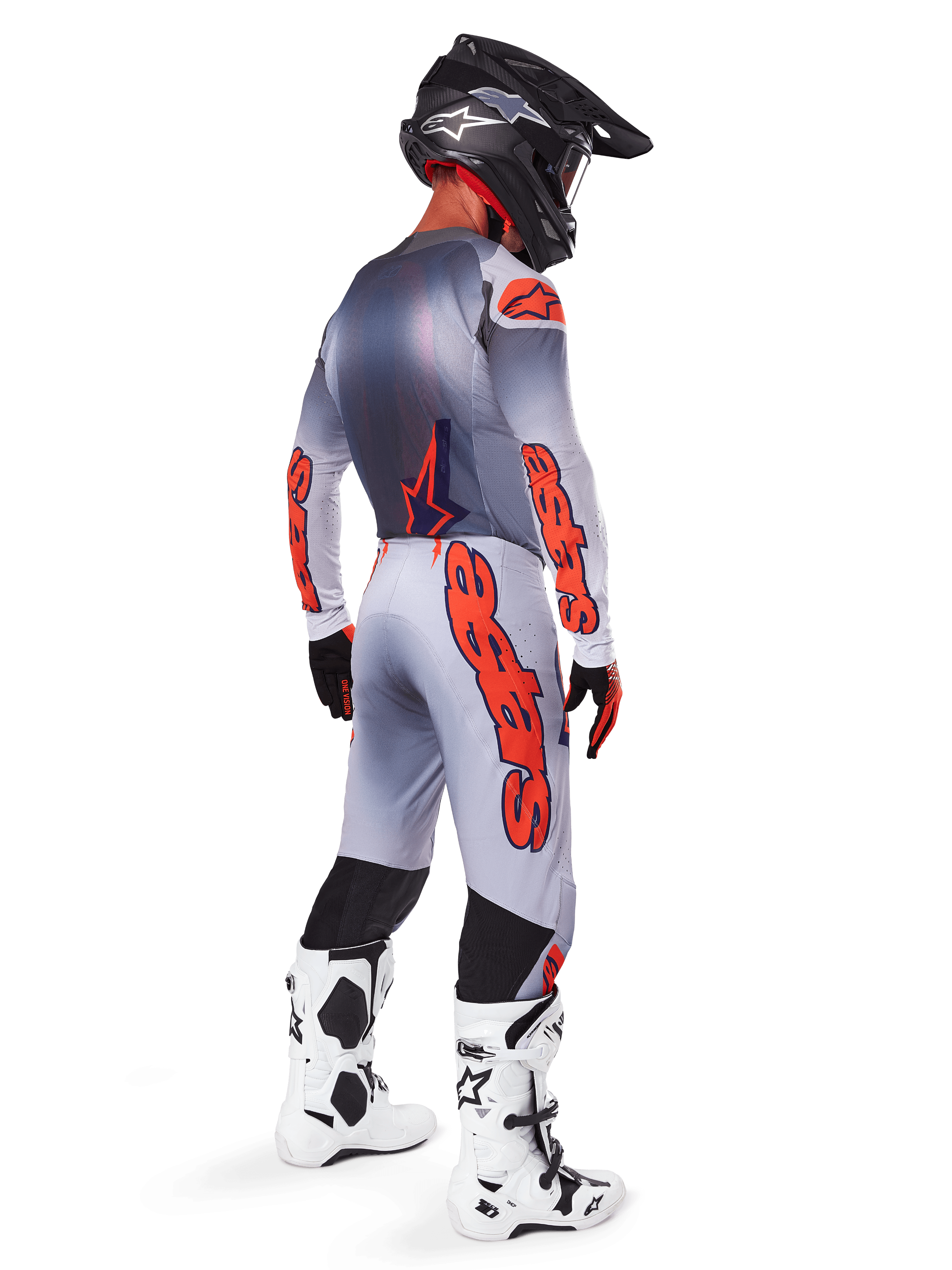 Piloto de motocross visto desde atrás vistiendo pantalones Alpinestars 2025 Supertech Lipan en gris y naranja flúor, jersey de manga larga a juego, casco negro y botas técnicas blancas y negras, equipamiento profesional completo de carreras off-road.