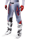 Pantalones Alpinestars Supertech Lipan 2025, pantalones de motocross, gris y naranja flúor, con diseños de estrellas azul marino y paneles interiores negros en las rodillas, usados con guantes rojo anaranjado y botas blancas, equipo profesional de carreras todoterreno