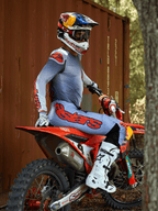 Piloto de motocross sentado en una moto de cross roja, vistiendo el conjunto Alpinestars 2025 Supertech Lipan en gris y naranja flúor, pantalones azul claro con un llamativo logotipo de Astars en naranja, jersey a juego, botas de motocross blancas y un casco de Red Bull, equipamiento profesional de competición coordinado