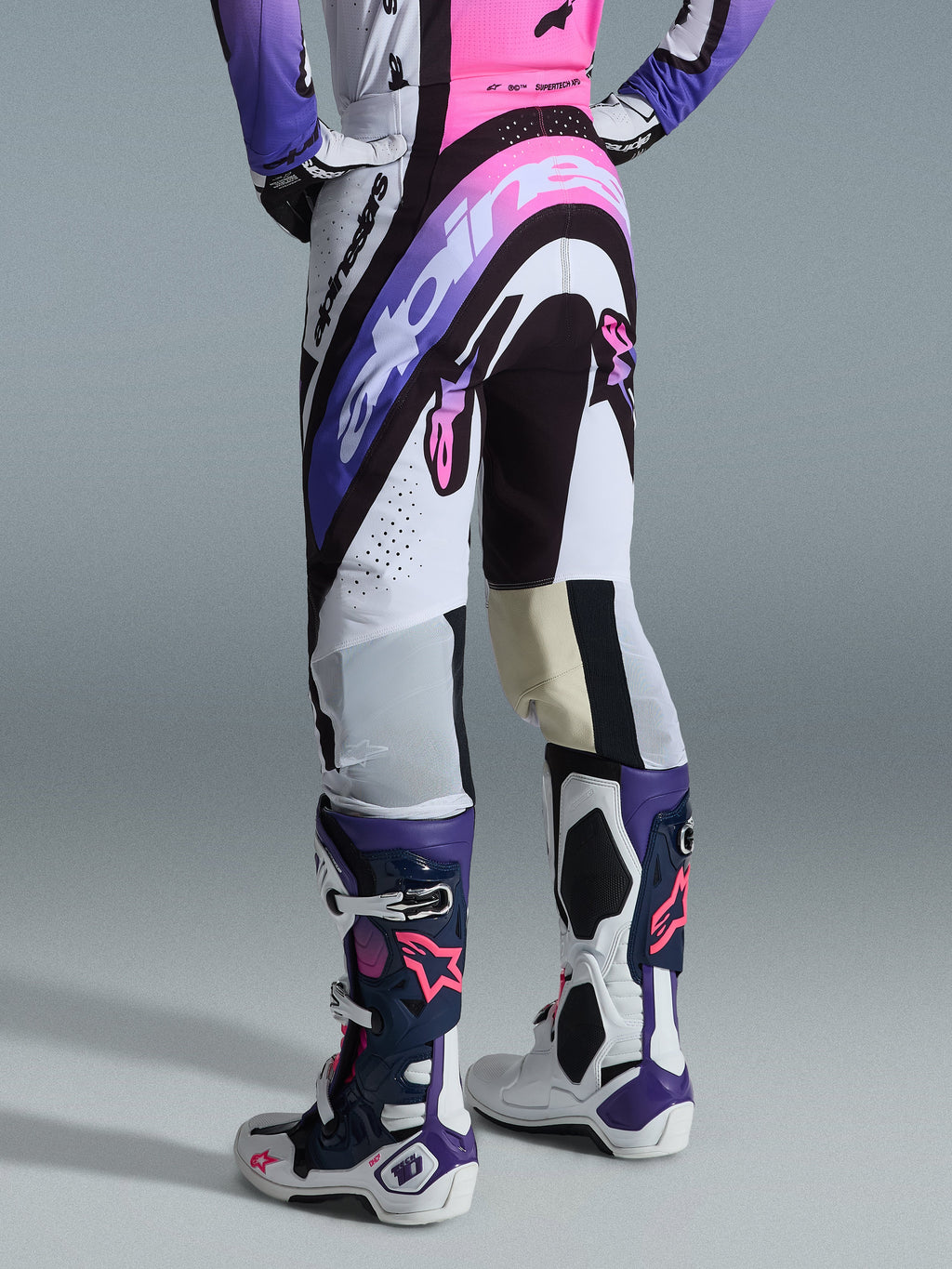 Alpinestars Supertech AFD Vista Pants 2026, pantalones de motocross, blancos, morados y negros con detalles rosas, vista trasera usados con jersey a juego y botas Tech 10, con logotipos llamativos y materiales de alto rendimiento para competición off-road de élite