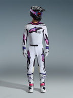 Piloto de motocross con un conjunto completo de equipamiento de competición, pantalones y jersey Alpinestars Supertech AFD Vista 2026 en blanco, morado y negro, guantes a juego, botas blancas y negras, casco negro con gafas moradas, ajuste atlético y estilizado para rendimiento off-road