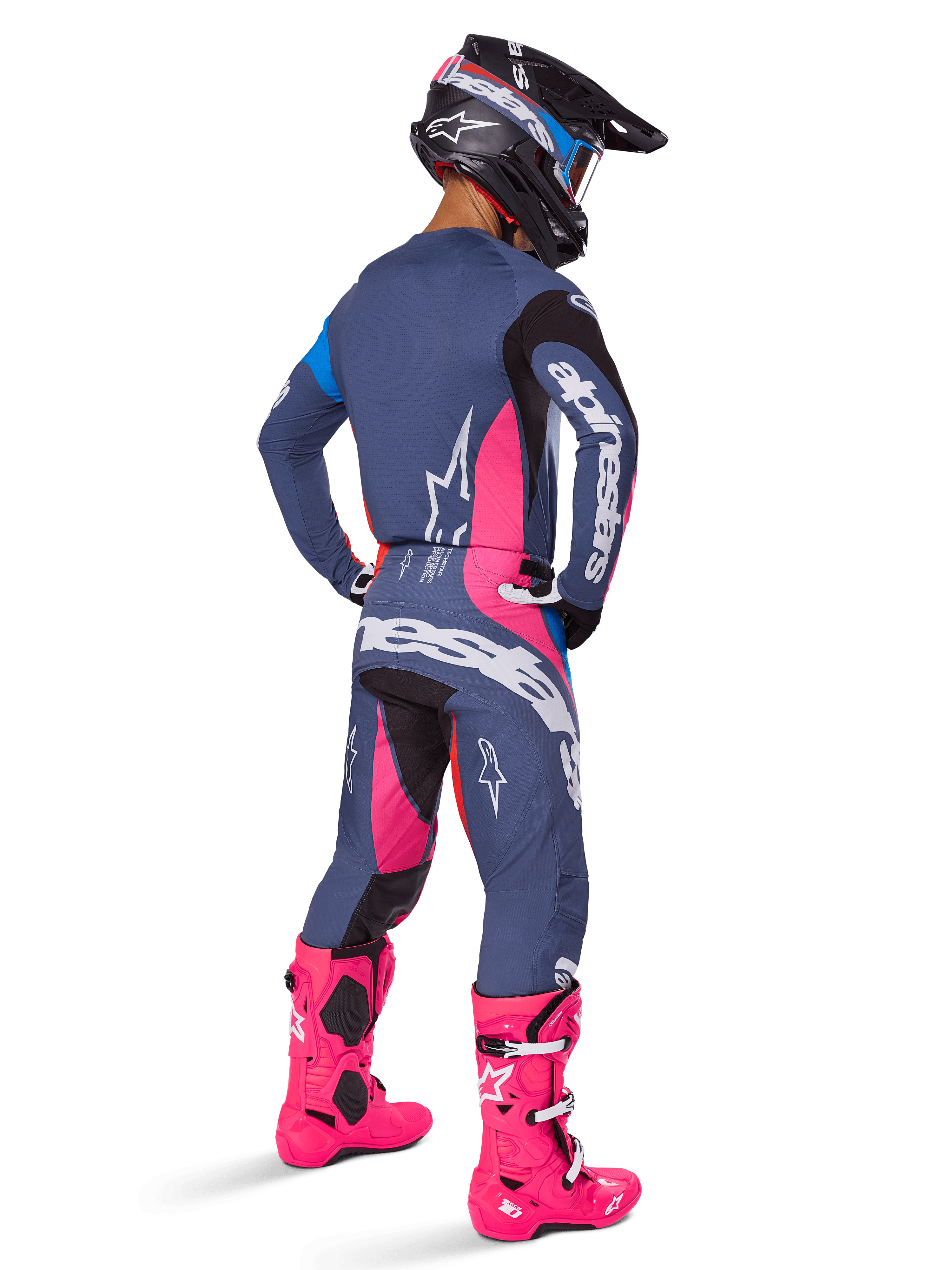 Piloto de motocross visto desde atrás con pantalones Alpinestars Techstar Dreem 2025, diseño en azul grisáceo y rosa magenta con detalles negros, combinado con jersey a juego y botas rosa vibrante, ajuste atlético de competición para rendimiento todoterreno