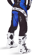 Pantalones Alpinestars Techstar Dreem 2025, pantalones de motocross para hombre, negros y azules, vista trasera con el llamativo logotipo blanco de Alpinestars en el asiento, combinados con botas de motocross blancas y negras y jersey azul a juego, ajuste atlético para competición off-road