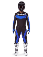 Piloto de motocross con el conjunto de equipo Alpinestars 2025 Techstar Dreem, camiseta negra con panel azul en el pecho y mangas, pantalones negros y azules a juego, casco negro con gafas y botas blancas, conjunto de motocross coordinado para carreras todoterreno.