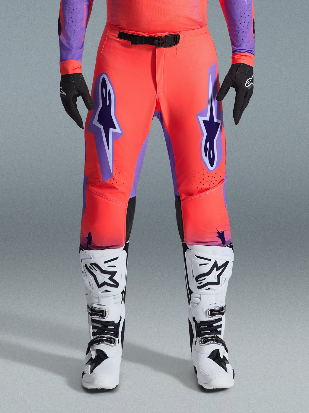 Alpinestars 2026 Supertech Scenz Pants, pantalones de motocross, en coral intenso y púrpura, combinados con jersey a juego, botas blancas y negras, y guantes negros, con grandes logotipos Astars púrpuras en los muslos y una hebilla magnética negra en la cintura, ajuste atlético profesional para competición