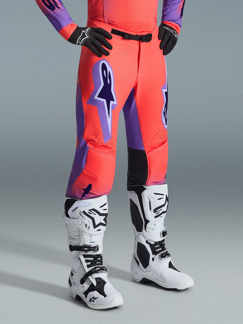 Pantalones Alpinestars Supertech Scenz 2026, pantalones de motocross, coral intenso y morado, usados con botas de motocross blancas y negras y guantes negros, con un gran logotipo morado de Alpinestars, paneles interiores de las rodillas en cuero negro y diseño de ajuste atlético para competición